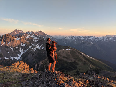 A man holds his dog while standing on the top of a mountain trail. 