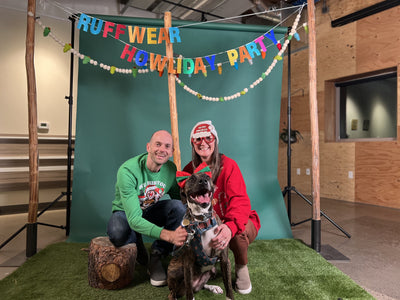 A woman, man, and their dog dressed in holiday outfits pose for a picture at the Ruffwear Howliday Party photo booth.