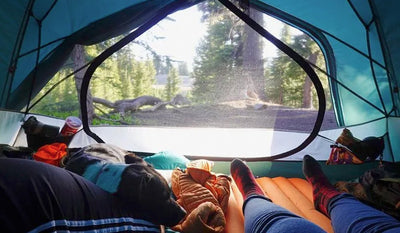 Two humans snuggled up with dog inside tent while camping.