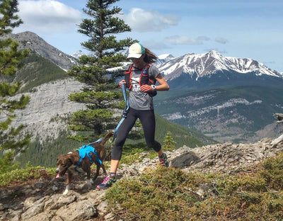 A woman runs on a mountain trail with her dog.  