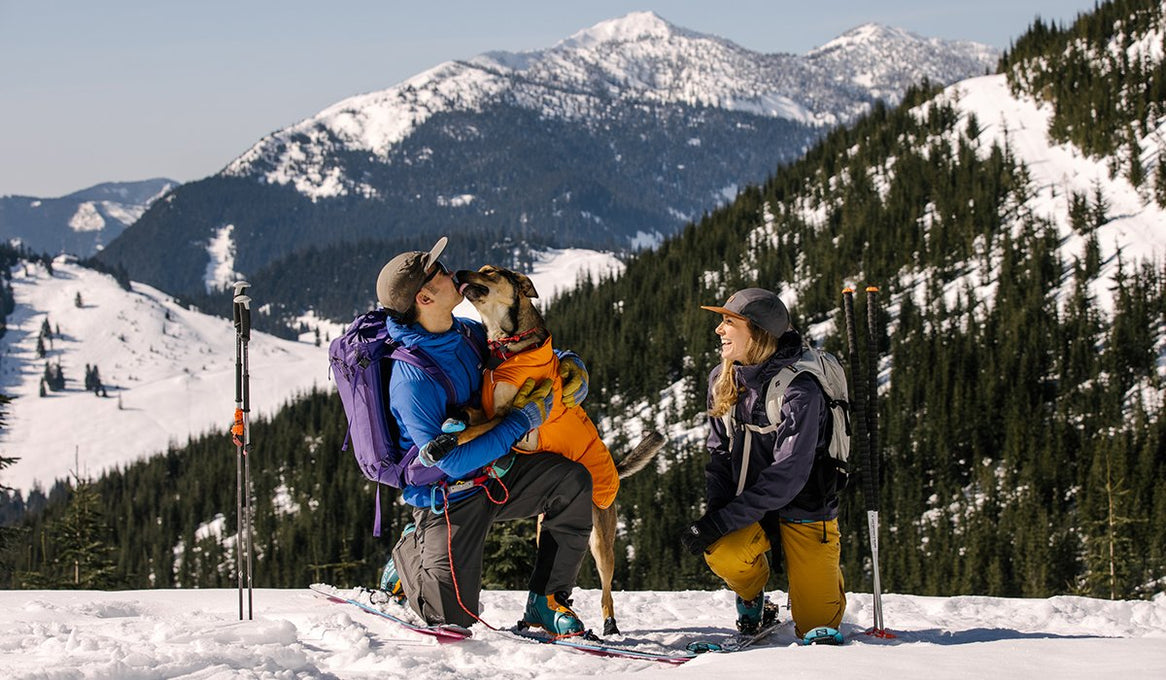 A dog and its two owners play in the snow with the Ruffwear Quinzee™ Dog Jacket. 