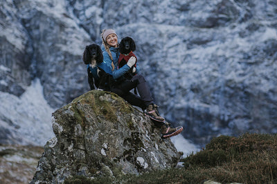 Two dogs wearing the Vert Jacket sit on a rock with their human in winter.