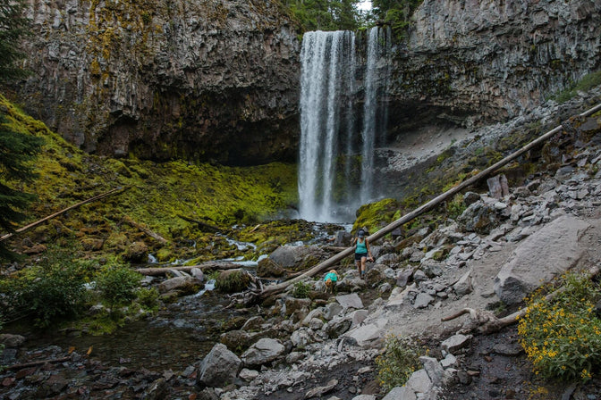 WOman and dog in matching teal pack and shirt walk down rocks in front of waterfall.